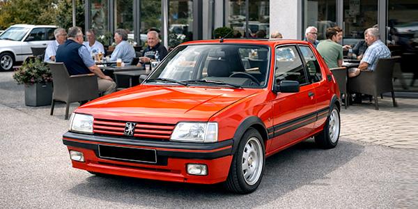 Peugeot 205 GTI als 80er-Hot-Hatch vor einem Cafe, wo sich Jung und Alt über Oldtimer, Fahrspaß und Gebrauchtwagen aus den 1980er-Jahren austauschen.